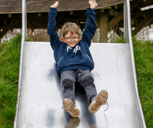 Boy with glasses in a blue hoodie is sliding down a slide with his arms raised in the air